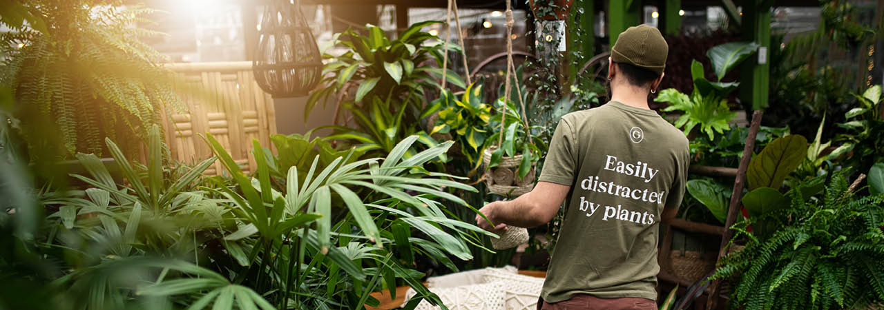 GARDENWORKS Employee wearing a t-shirt with "Easily Distracted by plants" on the back, immersed in a garden center with Tropical Plants 
